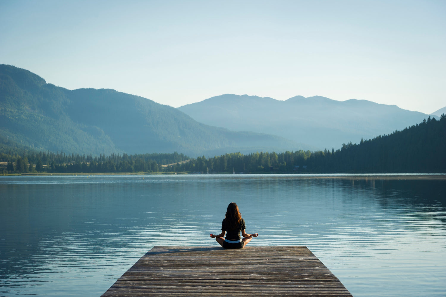 Person meditating on a dock by a lake with mountains in the background