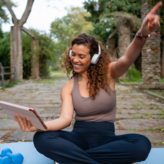 Woman sitting on a yoga mat outdoors, using a tablet and wearing headphones.